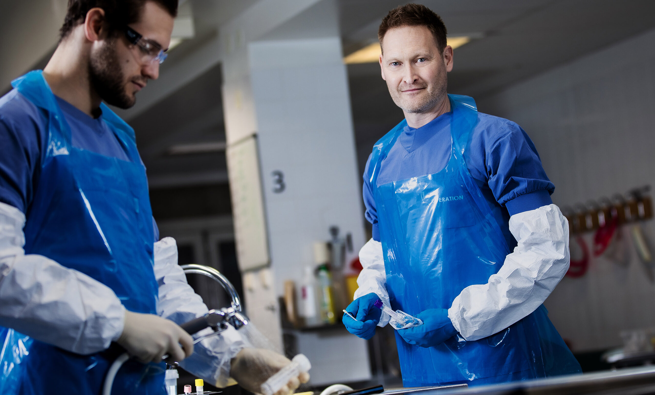 Två män med blå plastförkläden i obduktionssal