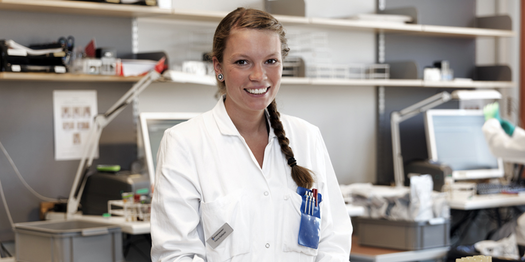 Woman wearing a lab coat standing in an office environment and smiling at the camera.